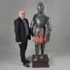 ARMADURA COMPLETA DE CAVALEIRO — TOLEDO, ESPANHA, SÉC. XIX - XX<br />Aço trabalhado e damasquinado | Altura: 1,80 m | Largura: 68 cm<br />Réplica historicista ou conjunto compósito de parada | Ótimo estado<br />Base em madeira nobre | Saiote têxtil vermelho e dourado<br /><br />Imponente armadura completa de cavaleiro, manufatura de Toledo, Espanha, em aço trabalhado e damasquinado. Elmo fechado tipo armet, peitoral globular, escarcelas, manopla, grevas e sapatos articulados. Montagem por ganchos, porcas e correias de couro com fivelas, conforme técnica dos séculos XV-XVI. Saiote em tecido vermelho com motivos dourados e franjas — traje de parada. Apresentada sobre plinto de madeira para exposição.<br /><br />A obra, a marca e a história<br />Tipologia: armadura de parada, revivalismo séc. XIX - XX<br /><br />As armaduras de placas surgiram no séc. XIV e atingiram a perfeição técnica no final do séc. XV. Em meados do séc. XVI, deixaram o campo de batalha e viraram vestido de gala para torneios e corte — ricamente cinzeladas, gravadas e damasquinadas por mestres de Milão, Augsburgo e Toledo.<br /><br />Esta peça, com 1,80 m, reproduz fielmente um arnês de campo e justa, c. 1550-1580, típico do reinado de Carlos V / Felipe II. Porém, a perfeição do aço, a ausência de marcas de uso e o saiote têxtil em ótimo estado indicam que se trata de réplica historicista do séc. XIX - XX — período Romântico em que Toledo retomou a produção para colecionadores, museus e castelos. O Castelo de Santa Catalina, Jaén, Andaluzia, possui exemplar idêntico em seu acervo, como citado.<br /><br />Toledo é mundialmente famosa desde o séc. XV pelo aço toledano, usado em espadas, armaduras e damasquinados. As Real Fábricas de Armas de Toledo, ativas até hoje, produziram nos sécs. XVIII-XIX milhares de armaduras de decoração para palácios europeus. A técnica de damasquinado — incrustação de ouro e prata em aço — é assinatura da cidade.<br /><br />Ótimo estado de conservação, sem oxidação ativa, indica peça de interior. Armaduras originais de combate são raríssimas e quase sempre fragmentadas. Conjuntos completos e íntegros são, em 95% dos casos, obras do Revivalismo Romântico, 1830-1910, feitas para a aristocracia e burguesia decorar halls, bibliotecas e castelos. A Real Armería de Madrid guarda o maior acervo original, com peças de Carlos I / V, e serviu de modelo para essas réplicas.<br /><br />Mercado e relevância<br />Armaduras completas historicistas de Toledo, séc. XIX, 1,80 m: € 8.000 a € 35.000 = R$ 48.000 a R$ 210.000. Se conjunto original séc. XVI documentado, ultrapassa € 500.000. Com damasquinado, base e saiote, em ótimo estado. No Brasil. Alta procura para decoração de alto padrão, hotéis, vinícolas e coleções de militaria.<br /><br />É Toledo em aço: cavaleiro, damasquino e honra. A armadura que guardou salões do Romantismo — 1550 ou 1880 forjada em 1,80 m, reluzindo em 2026.