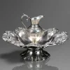 GOMIL E BACIA — EDWARD BARNARD & SONS<br />Londres, 1851 | Prata Sterling .950 | Período Vitoriano<br />Bacia: 12 x 33 x 33 cm | Jarro: 17 x 13 x 10 cm | Peso: 1.106 kg<br /><br />Monumental gomil e bacia em prata Sterling, Edward Barnard & Sons, Londres, 1851. Conjunto para abluções — lavar as mãos antes de refeições ou cerimônias — forma que remonta à idade média e atinge o ápice no séc. XIX vitoriano.<br /><br />As marcas: Garantia inglesa completa<br />A) Leão passante — Lion Passant: garantia da prata Sterling .925+, aqui .950, padrão superior.<br />B) Leopardo coroado — marca do Assay Office de Londres, ativo 1478–1821 para prata e retomado para ouro.<br />C) Letra data “Q” — Date Letter: 1851, reinado de Victoria.<br />D) Cabeça da Rainha Victoria — Duty Mark: imposto pago sobre prata entre 1784–1890.<br />E) Marca do fabricante: EB & S — Edward Barnard & Sons, casa fundada 1680, fornecedora da Corona Britânica. Criaram a célebre “Lily Font”, pia batismal em prata dourada usada nos batismos reais desde 1841.<br />Remarcação: C. Taylor & Son — Bristol: casa comercial revendedora, prática comum no séc. XIX.<br /><br />Bacia: bordo polilobado com galeria vazada em parras, uvas e arabescos — Rococo Revival. Fundo martelado ao centro com brasão vitoriano gravado. Base em pé de cálice gomado.<br /><br />Jarro: corpo ovoide com guilhochê, cartelas e caneluras. Bico aberto, alça em voluta folheada. Todo o conjunto em 1,106 kg de prata maciça de espessura e peso relevantes.<br /><br />A relevância: Edward Barnard & Sons<br />Ativa por 300 anos, a Barnard foi a maior ourivesaria de Londres. Além da Lily Font — usada até hoje nos batismos de George, Charlotte e Louis de Cambridge — produziu as joias da Coroa menores e baixelas para a nobreza. Peças de 1851 coincidem com a Grande Exposição de Londres, ápice do design vitoriano.<br /><br />Mercado<br />Gomil e bacia Barnard & Sons, 1851, com marcas completas e brasão: £ 4.000 a £ 15.000 = R$ 28.000 a R$ 105.000 em leilões internacionais. A qualidade do cinzelado, o peso acima de 1 kg e a procedência real colocam este conjunto no topo do mercado de prataria inglesa.<br /><br />É Londres em prata: leão, leopardo e Victoria. O ritual vitoriano que lavou mãos de duques — 1851 vertido em 1.106 gramas, reluzindo em 2026.